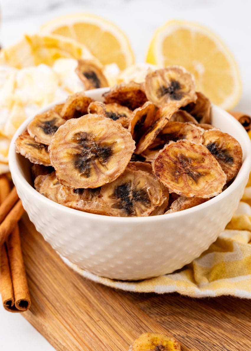 homemade banana chips in a small white bowl on a wooden cutting board