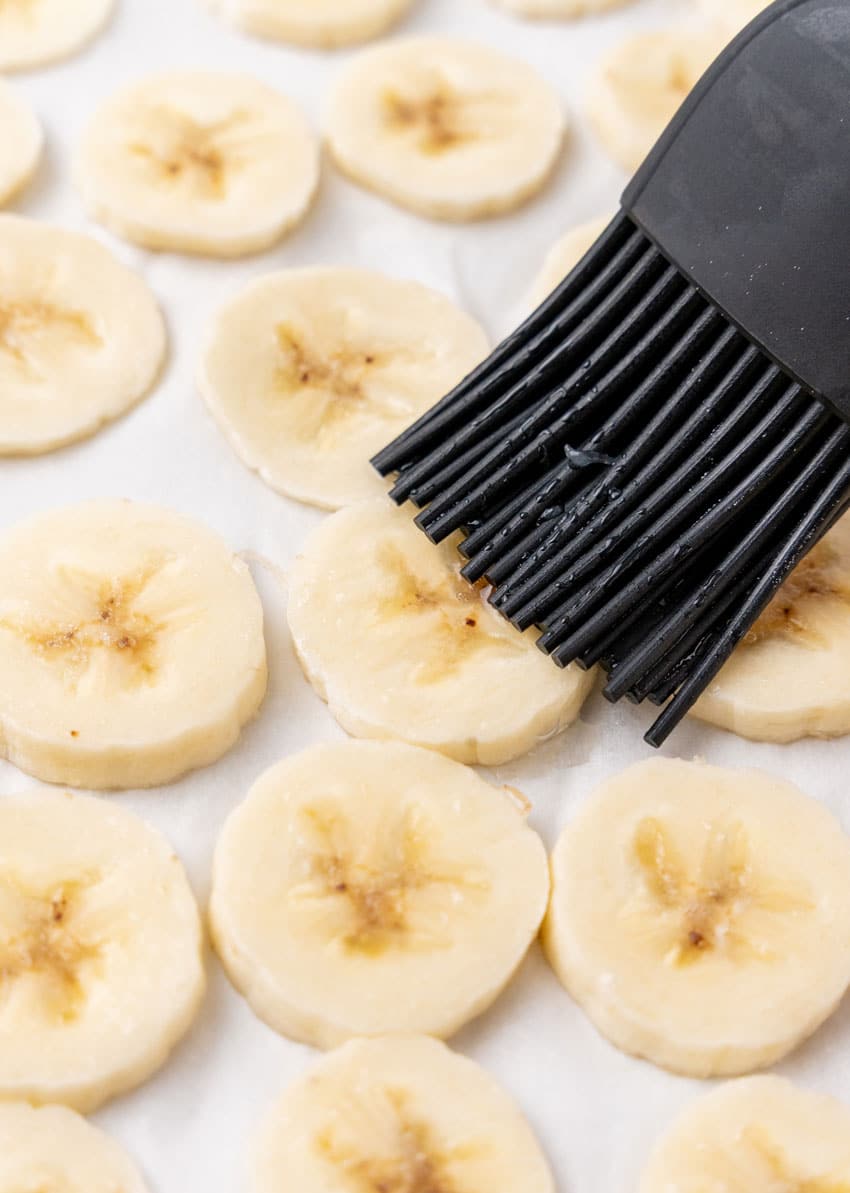 a silicone brush wiping lemon juice over fresh banana slices on parchment paper