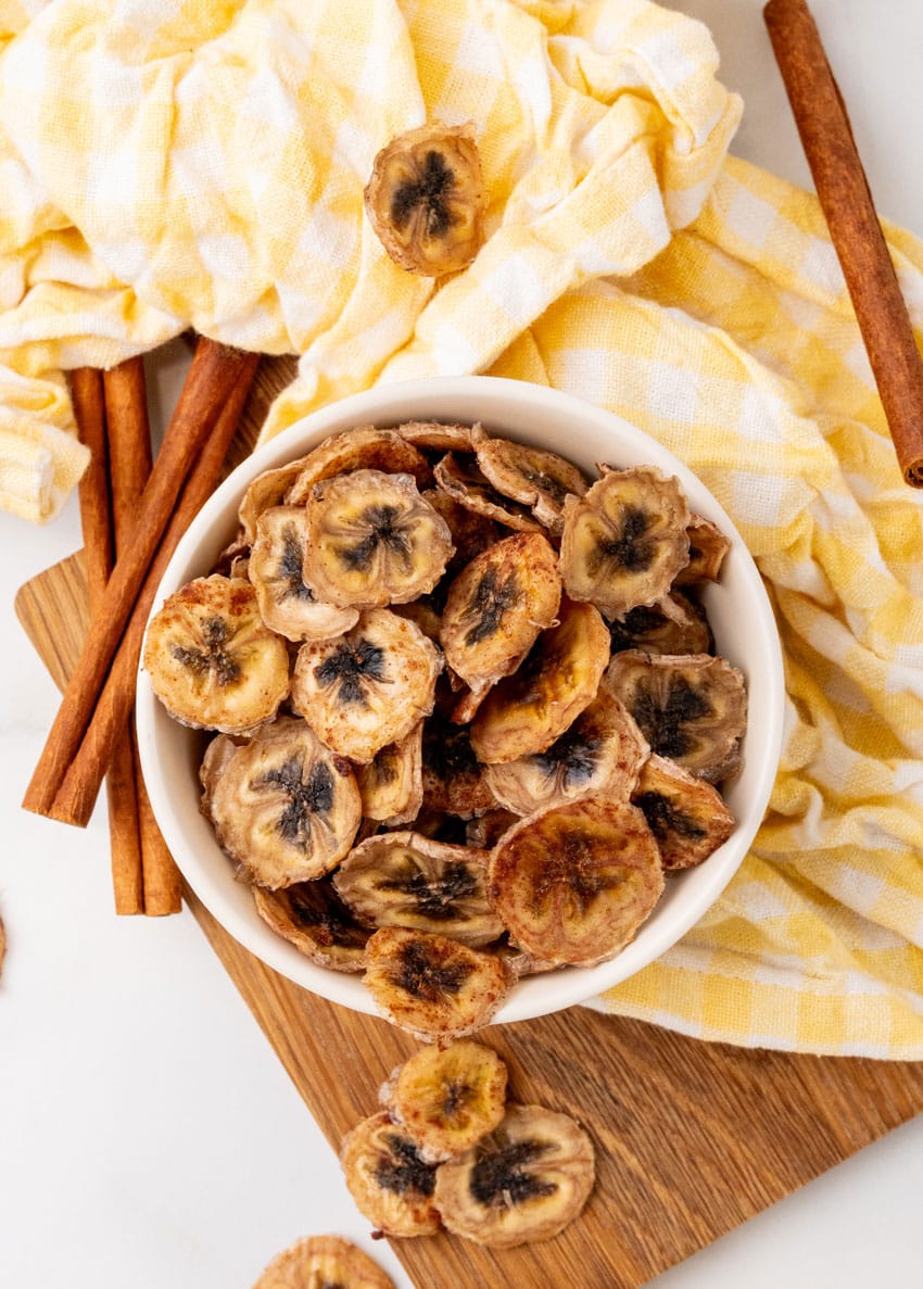 homemade banana chips in a small white bowl on a wooden cutting board
