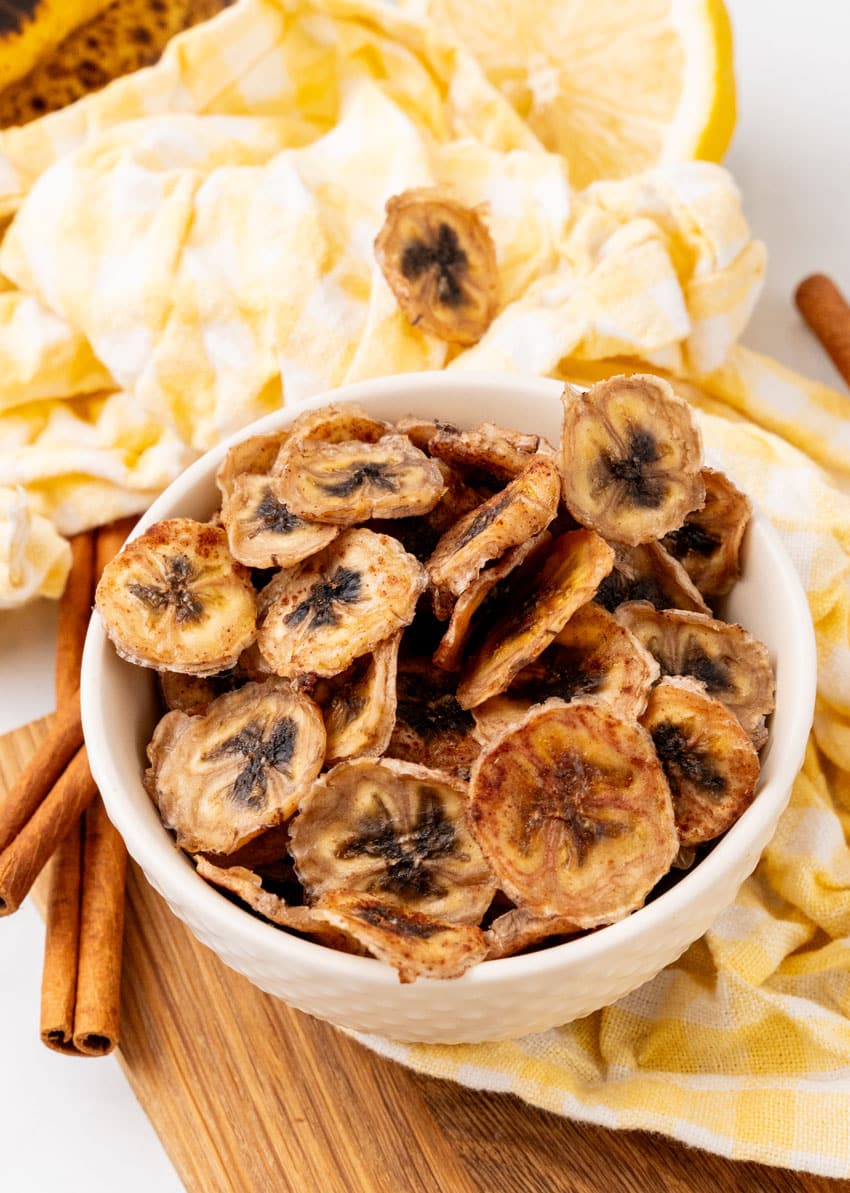 homemade banana chips in a small white bowl on a wooden cutting board
