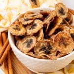 homemade banana chips in a small white bowl on a wooden cutting board
