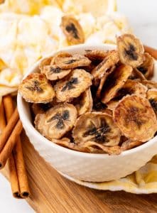 homemade banana chips in a small white bowl on a wooden cutting board