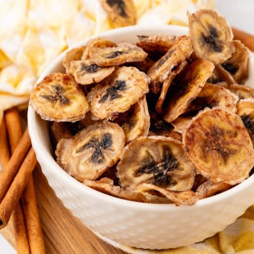 homemade banana chips in a small white bowl on a wooden cutting board