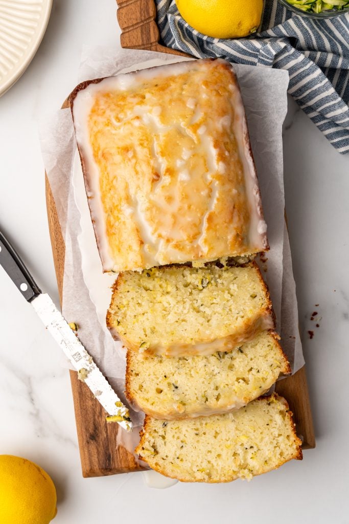 a sliced loaf of glazed lemon zucchini bread on a parchment covered wooden cutting board