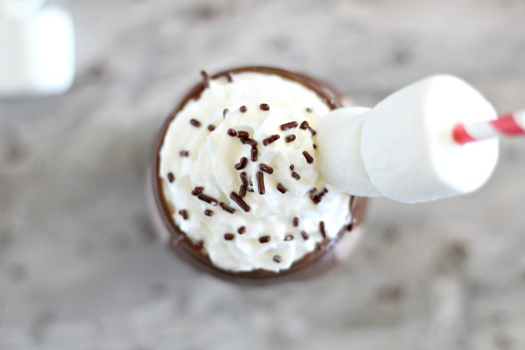 frozen hot chocolate shown topped with whipped cream, chocolate sprinkles, and a marshmallow topped paper straw