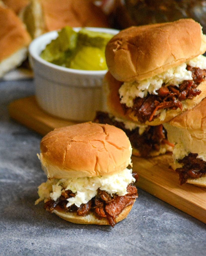 pulled pork sliders shown on a gray background with pickles and fresh rolls in the background
