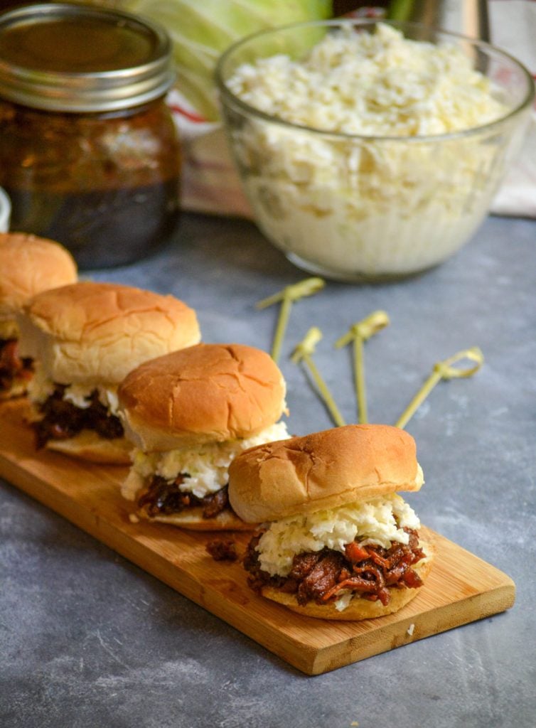 several balsamic & honey pulled pork sliders served on a narrow wooden cutting board with coleslaw and extra barbecue sauce in the background