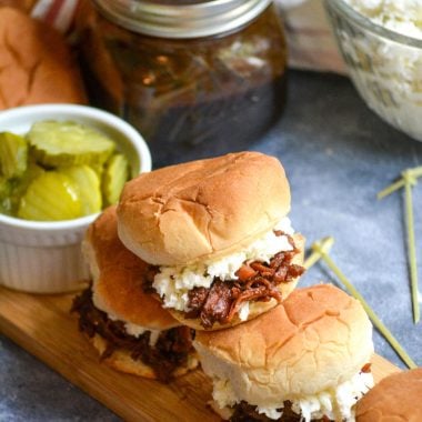 a stack of pulled pork sliders on a narrow wooden cutting board served with crisp dill pickle slices