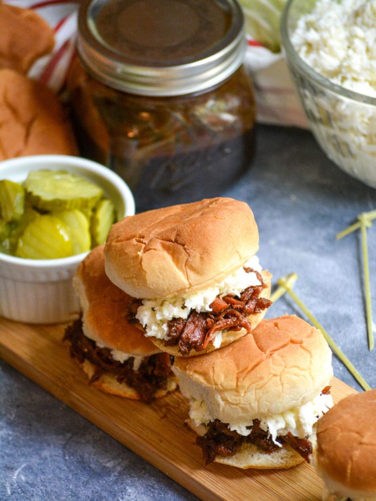 a stack of pulled pork sliders on a narrow wooden cutting board served with crisp dill pickle slices