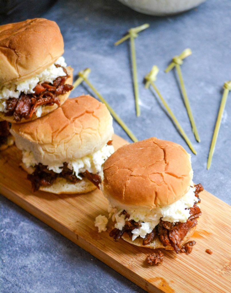 pulled pork slider sandwiches shown on a narrow wooden cutting board on a gray background