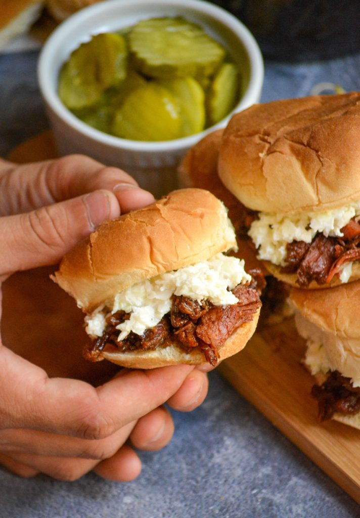 a hand holding up a coleslaw and pickle topped pulled pork slider