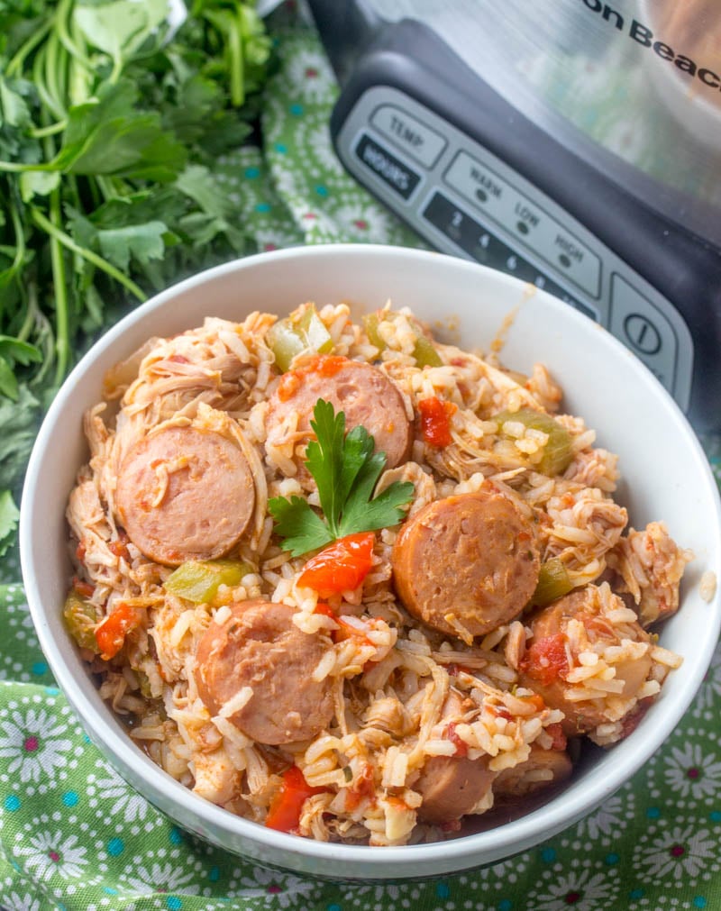 Crockpot Chicken, Sausage, & Rice shown in a white bowl with fresh herbs in the back ground