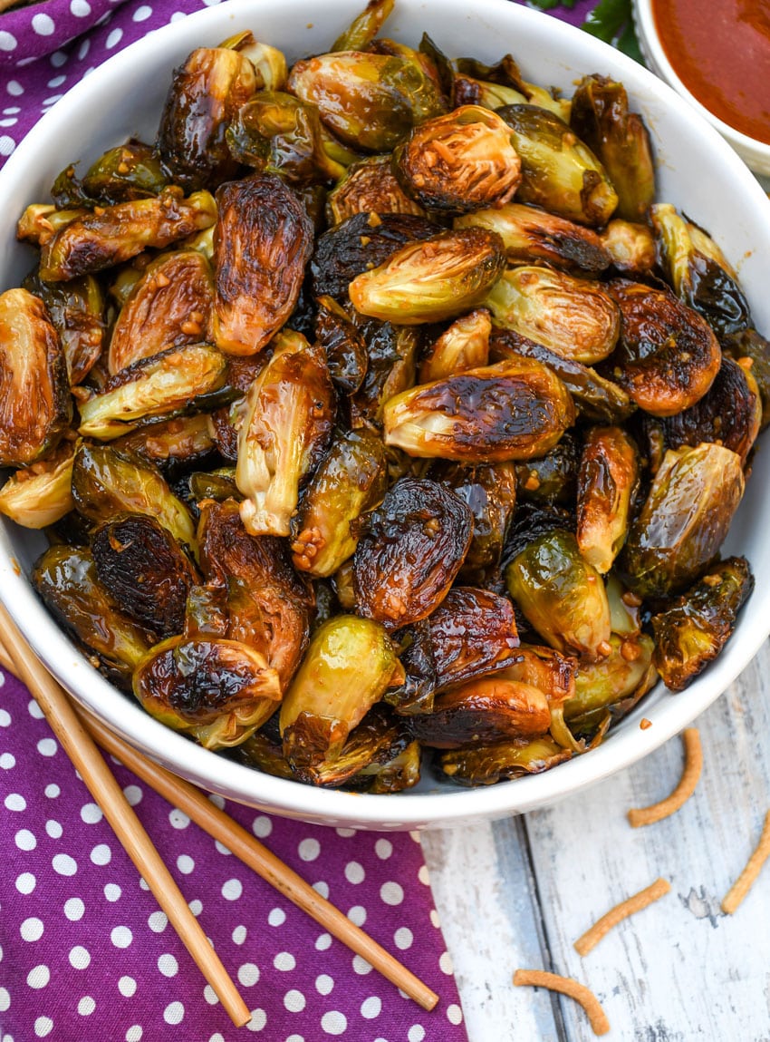 crispy soy glazed brussels sprouts in a white serving bowl with wooden chop sticks on the side