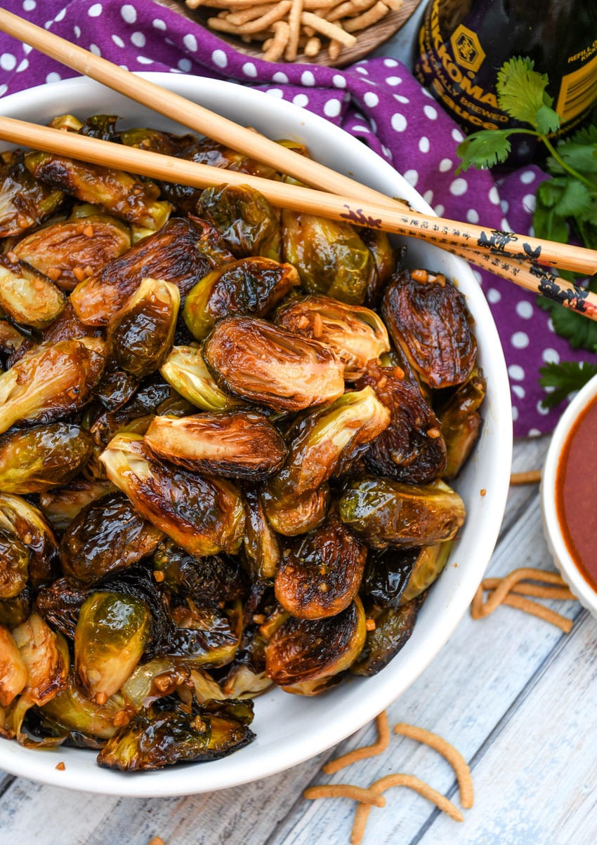 crispy soy glazed brussels sprouts in a white serving bowl with wooden chop sticks on the side