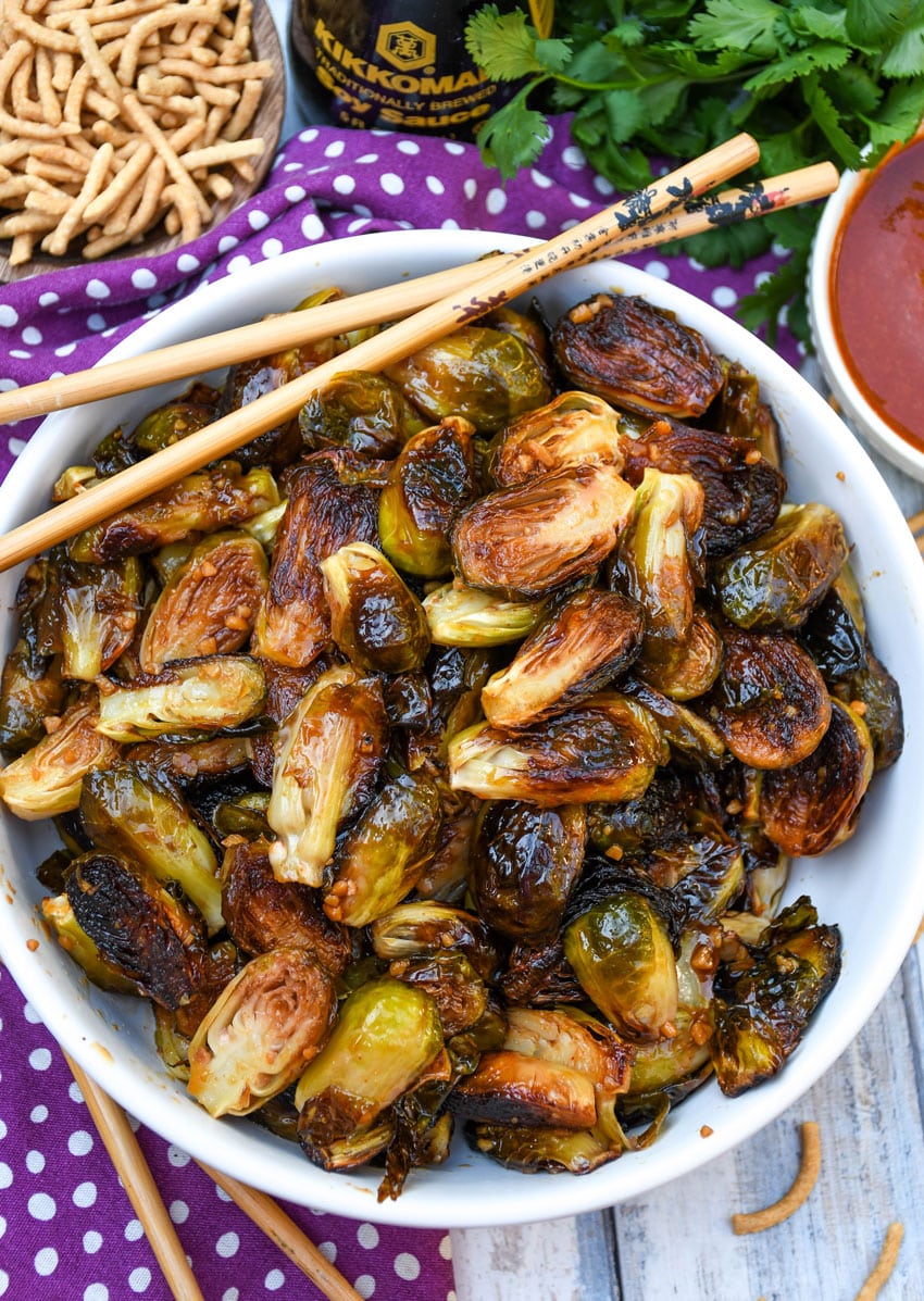 crispy soy glazed brussels sprouts in a white serving bowl with wooden chop sticks on the side