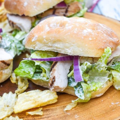 grilled chicken caesar sandwiches surrounded by potato chips on a wooden cutting board
