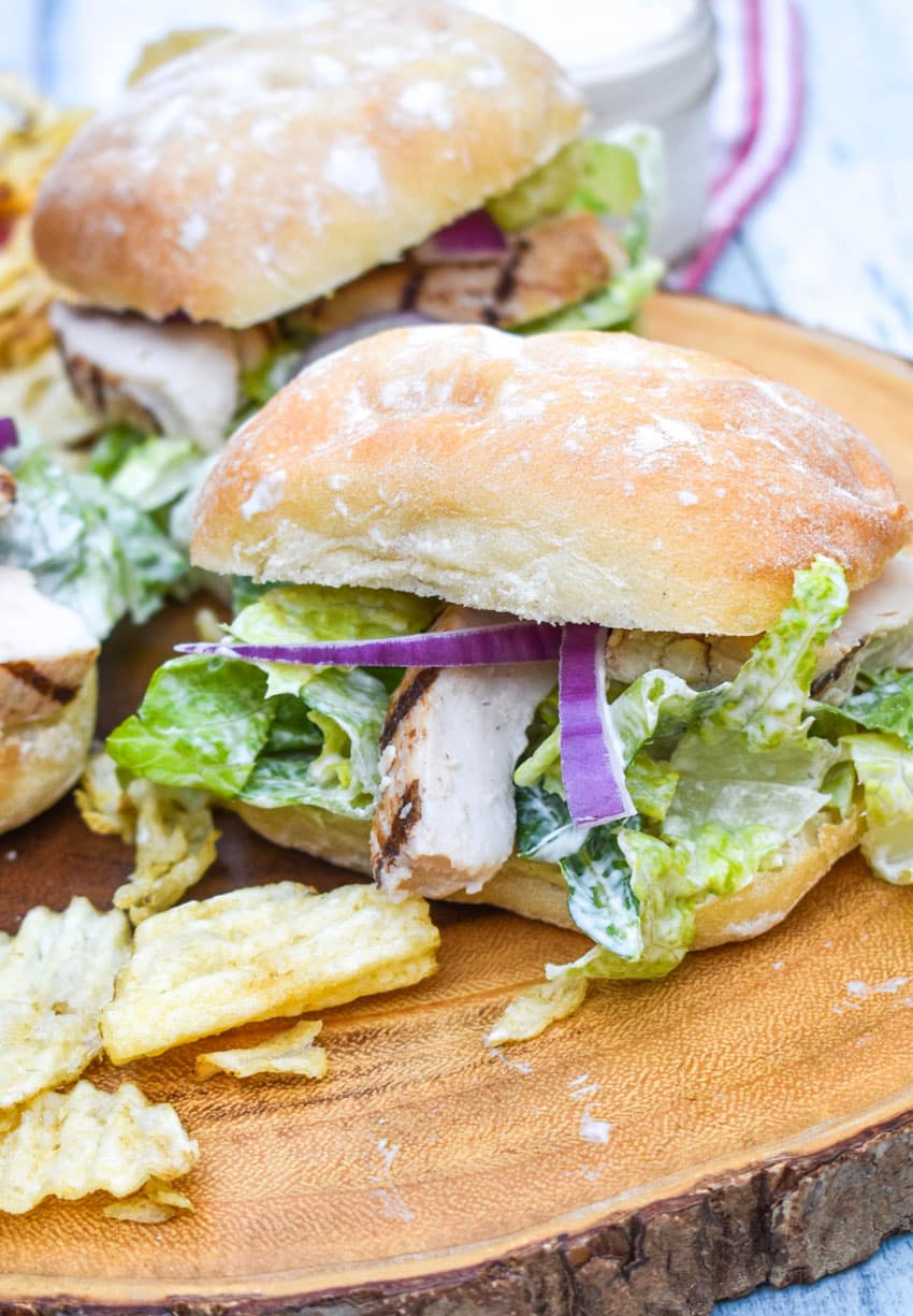 grilled chicken caesar sandwiches surrounded by potato chips on a wooden cutting board