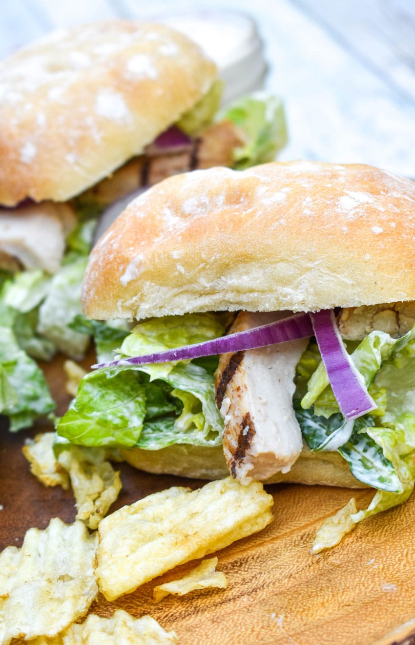 grilled chicken caesar sandwiches surrounded by potato chips on a wooden cutting board