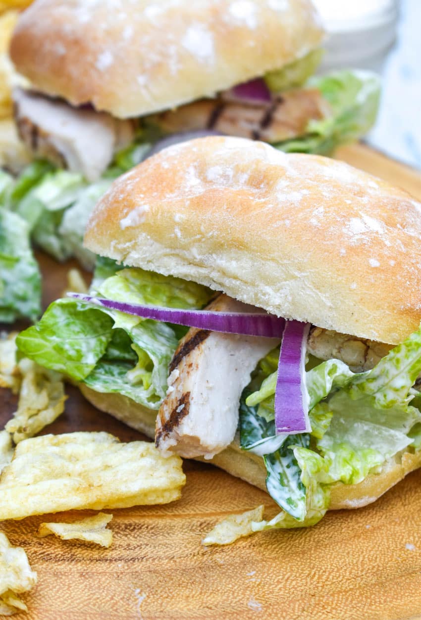 grilled chicken caesar sandwiches surrounded by potato chips on a wooden cutting board