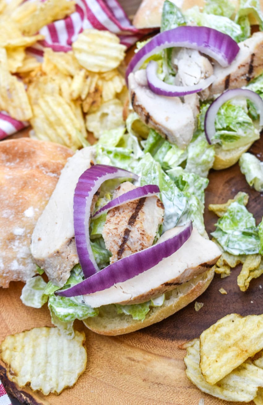 open grilled chicken caesar sandwiches surrounded by potato chips on a wooden cutting board