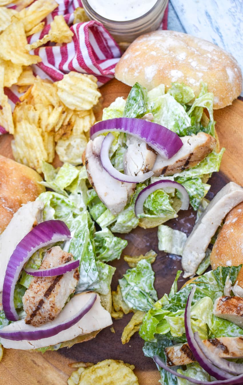 open grilled chicken caesar sandwiches surrounded by potato chips on a wooden cutting board