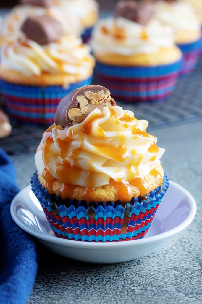 Baby Ruth Cupcakes shown on a wire cooling wrap with one on a small white plate in the foreground