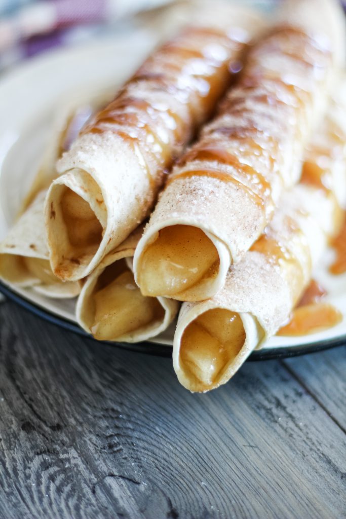 cinnamon sugar coated apple pie taquitos are shown stacked on a white place and drizzled with caramel syrup