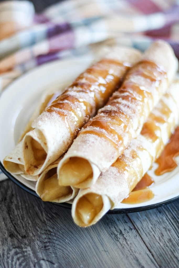 cinnamon sugar coated apple pie taquitos are shown stacked on a white place and drizzled with caramel syrup