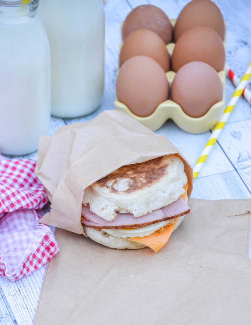 a freezer breakfast sandwich wrapped in a brown paper sandwich bag in front of brown eggs and two glasses of milk