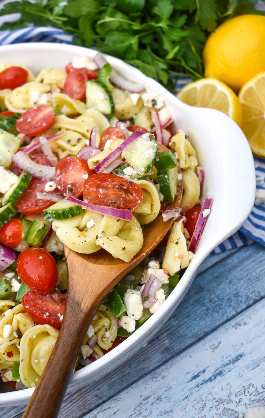 a wooden spoon scooping Mediterranean tortellini pasta salad out of a white serving bowl