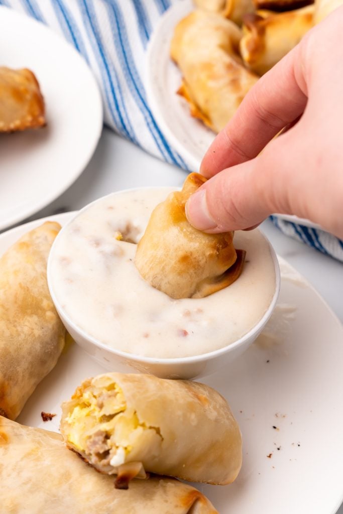 a crispy baked breakfast egg roll being dunked in a dipping bowl of homemade sausage gravy