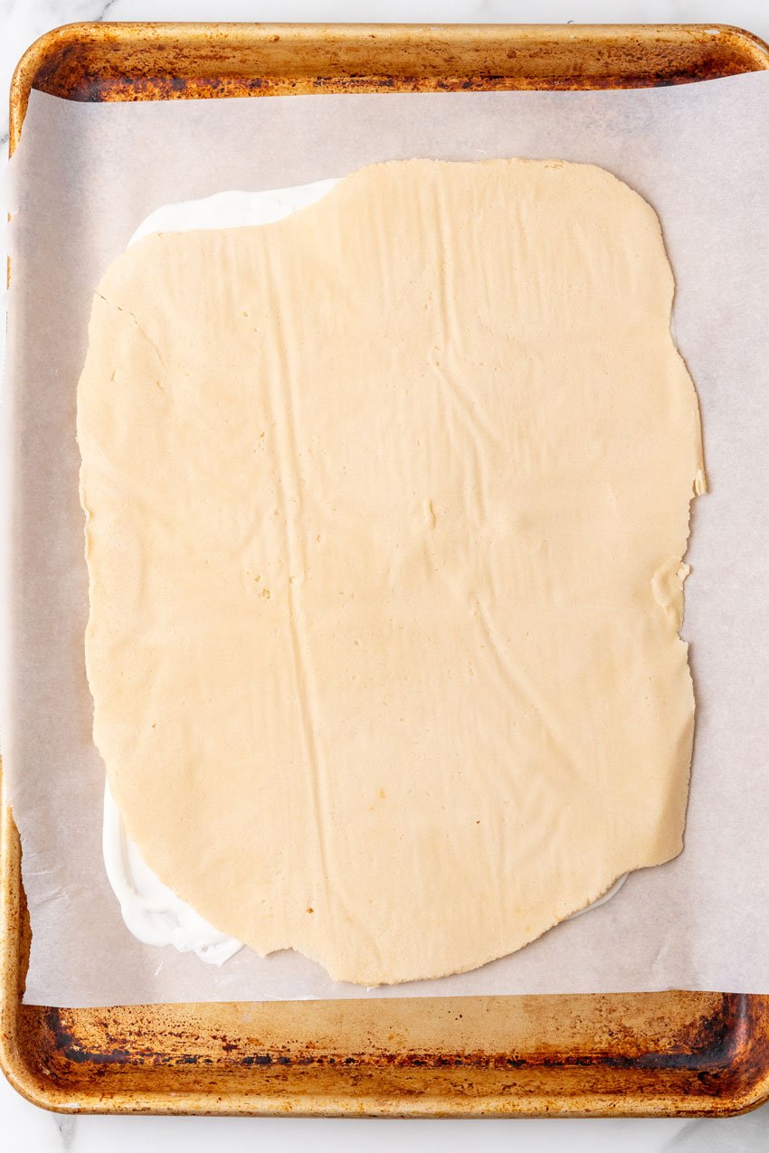 sugar cookie dough rolled out flat over a rectangle of melted white chocolate on a parchment paper lined baking sheet