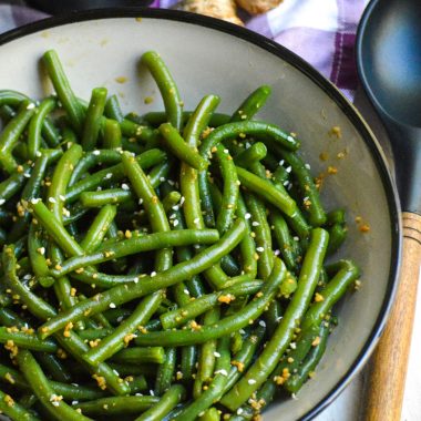 garlic ginger green beans served in a ceramic bowl with a wooden spoon, ginger root, and soy sauce in the background