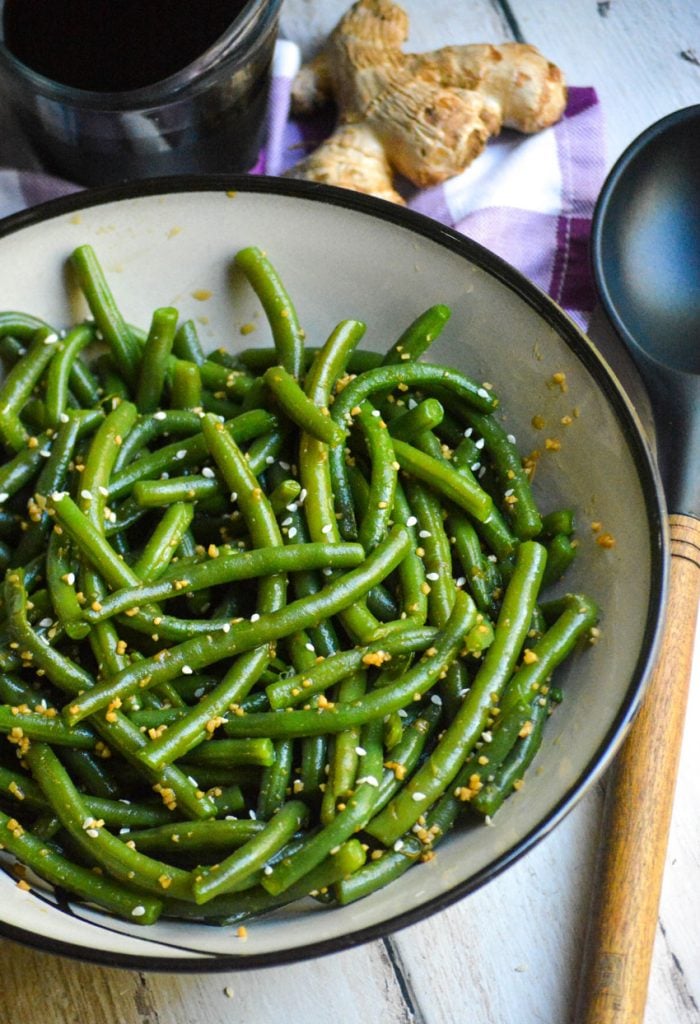 garlic ginger green beans served in a ceramic bowl with a wooden spoon, ginger root, and soy sauce in the background