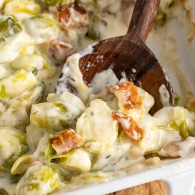 a wooden spoon stuck in brussels sprouts gratin in a white casserole dish