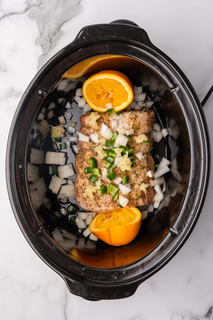 pork loin topped with diced veggies and orange halves in a black crockpot