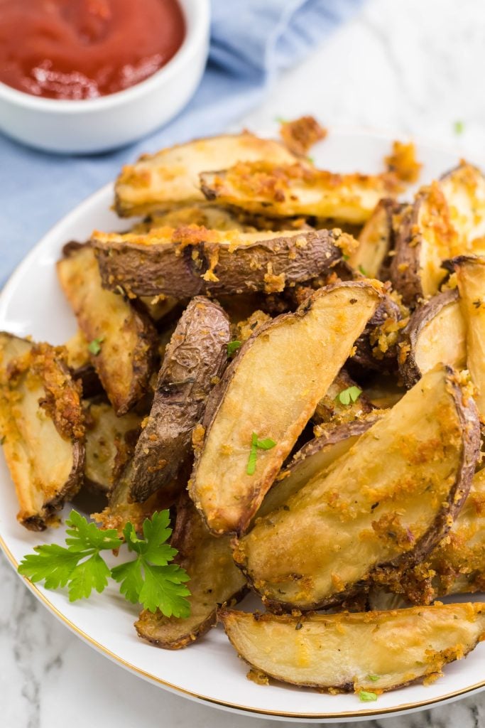 crispy oven baked cheese fries piled high on a white plate with parsley leaves for garnish and a bowl of ketchup in the background