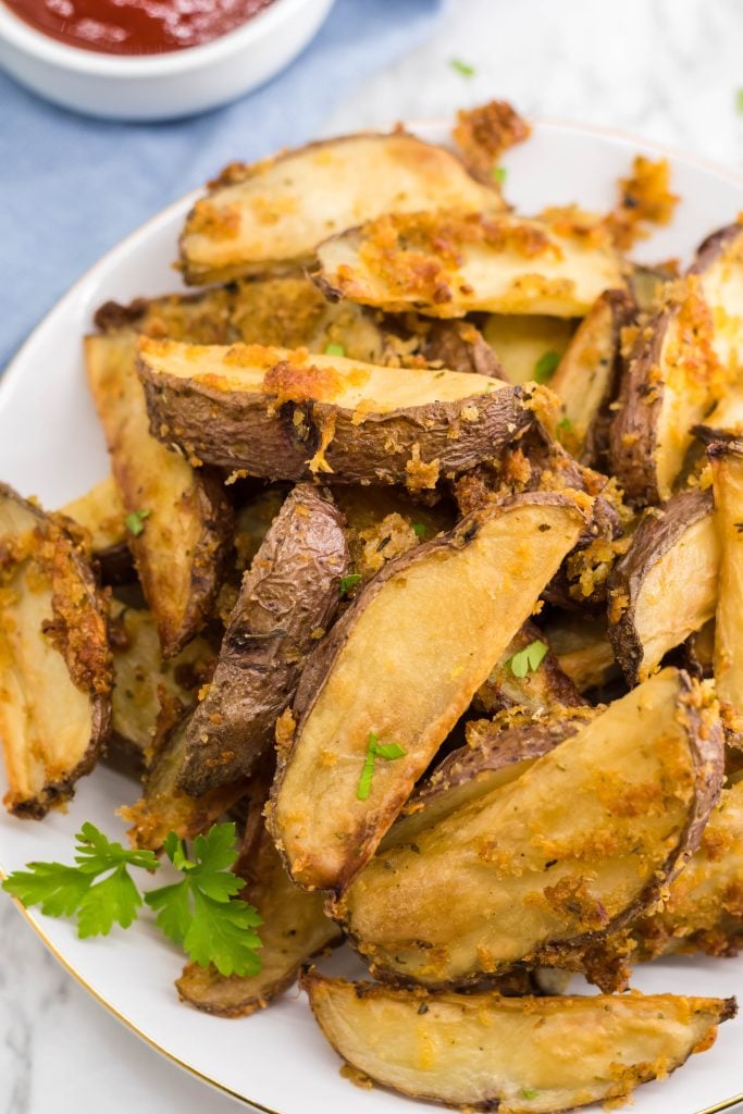 crispy oven baked cheese fries piled high on a white plate with parsley leaves for garnish and a bowl of ketchup in the background