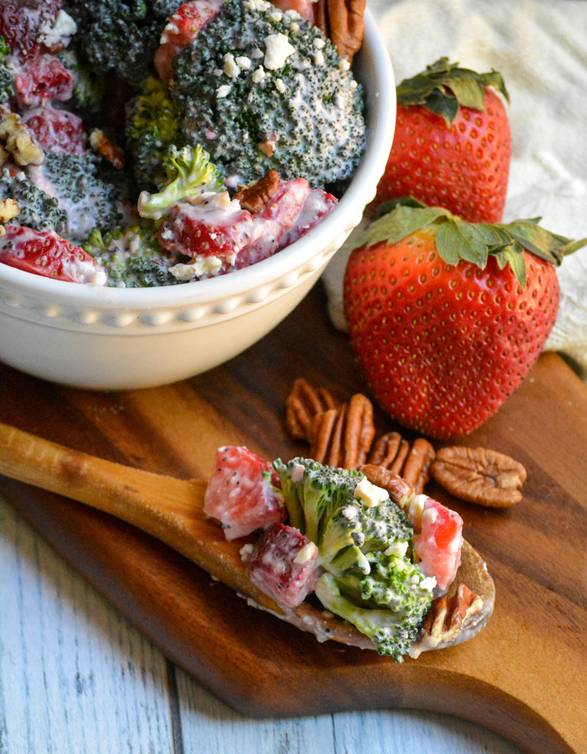 a wooden spoon topped with strawberry broccoli salad resting on a wooden cutting board beside nuts and fresh berries