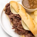 a crockpot french dip sandwich on a white plate with potato chips and small bowl of au jus on the side