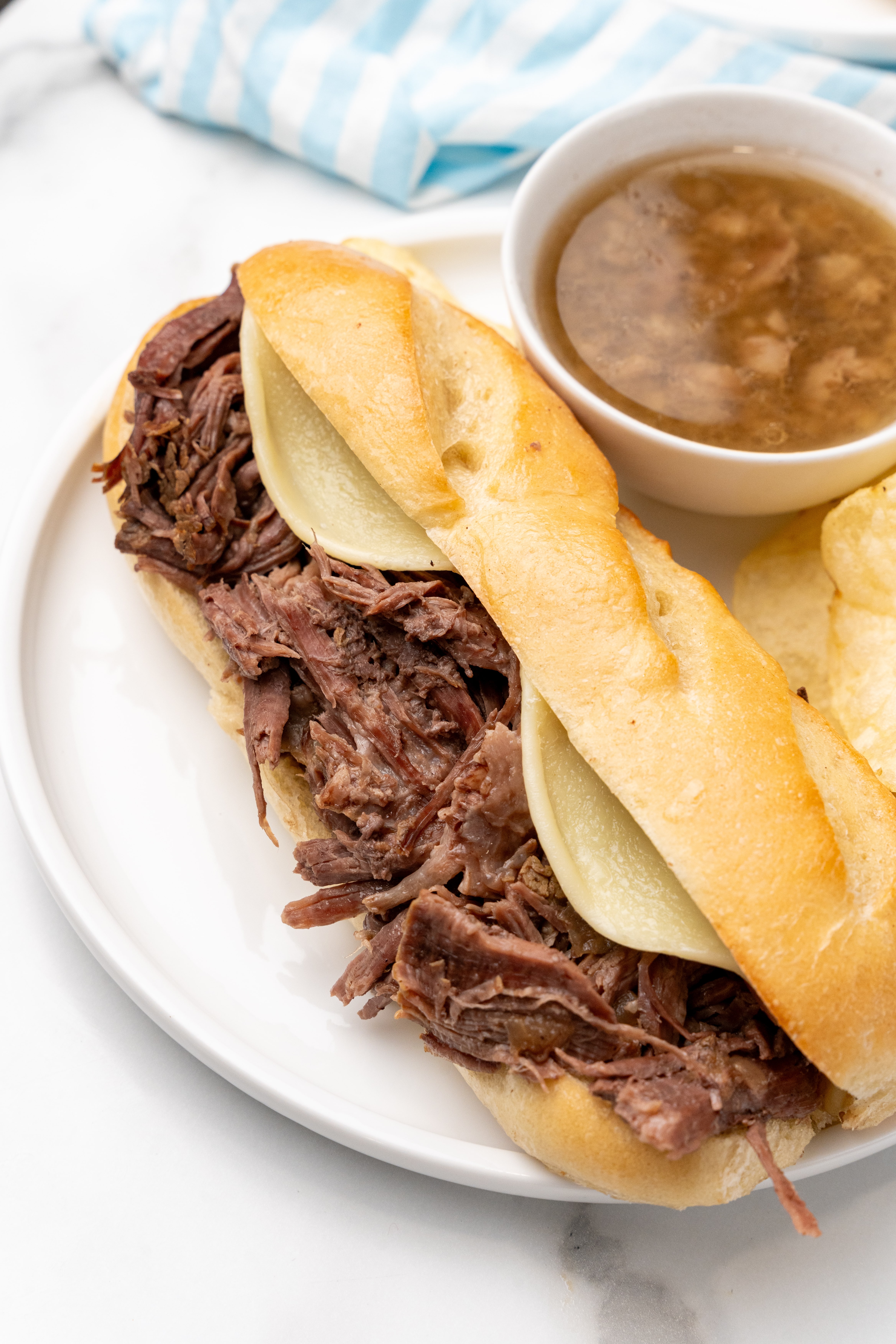 a crockpot french dip sandwich on a white plate with potato chips and small bowl of au jus on the side