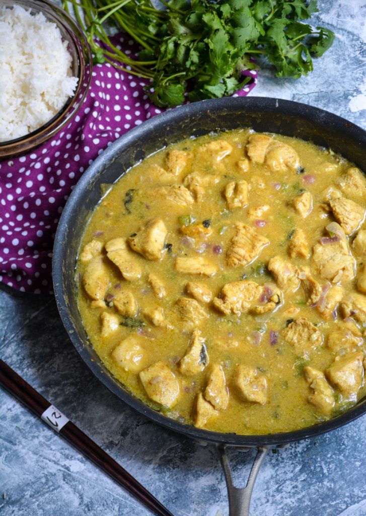 basil chicken in coconut curry sauce in a black skillet on a blue background with cilantro and white rice in the background