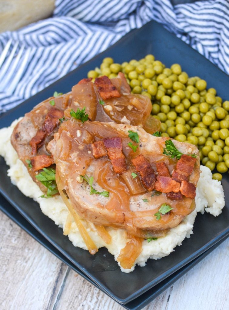 two smothered pork chops served over mashed potatoes with peas on the side on a black square plate