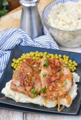 two smothered pork chops served over mashed potatoes with peas on the side on a black square plate