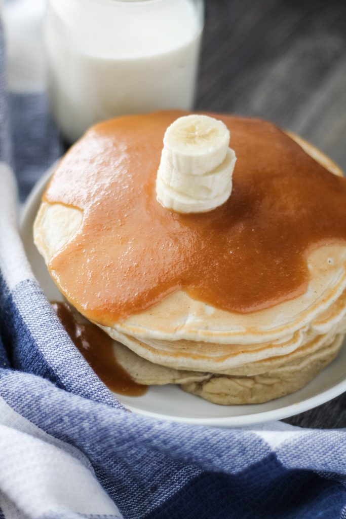 a stack of banana pancakes with peanut butter syrup shown on a white plate with a glass of milk in the background