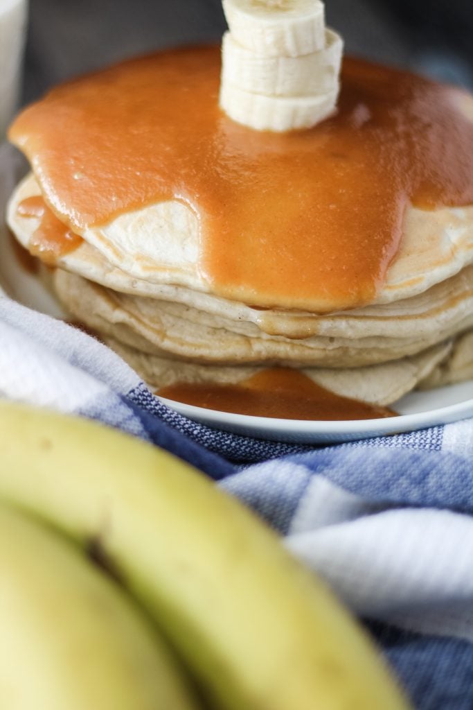 a stack of banana pancakes with peanut butter syrup shown on a white plate with a glass of milk in the background