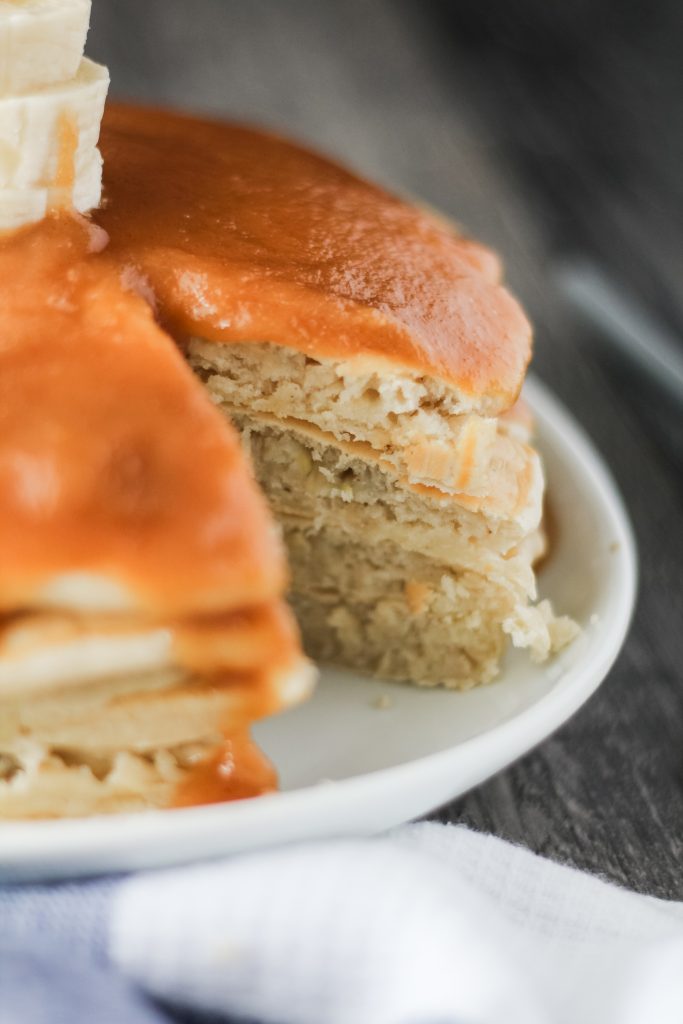 a stack of banana pancakes with peanut butter syrup shown on a white plate with a slice removed