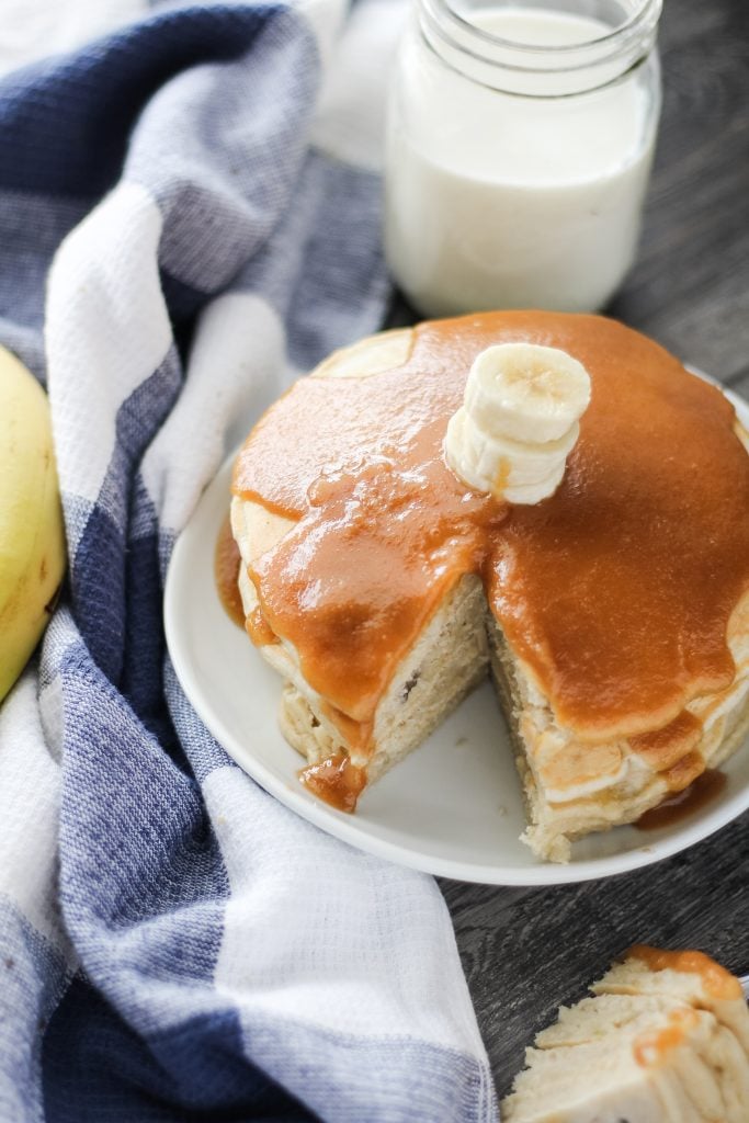 a stack of banana pancakes with peanut butter syrup shown on a white plate with a slice removed