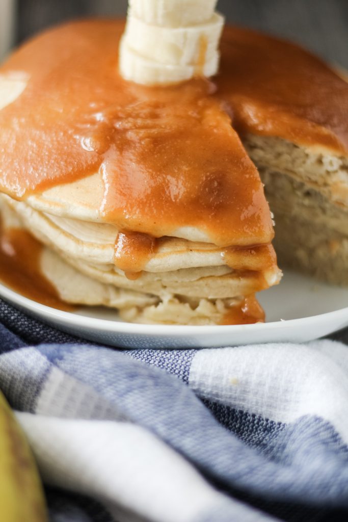 a stack of banana pancakes with peanut butter syrup shown on a white plate with a slice removed