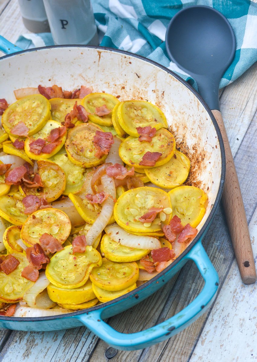 a wooden handled spoon resting next to a blue skillet filled with tender yellow squash slices studded with strips of onnion and crispy bits of bacon